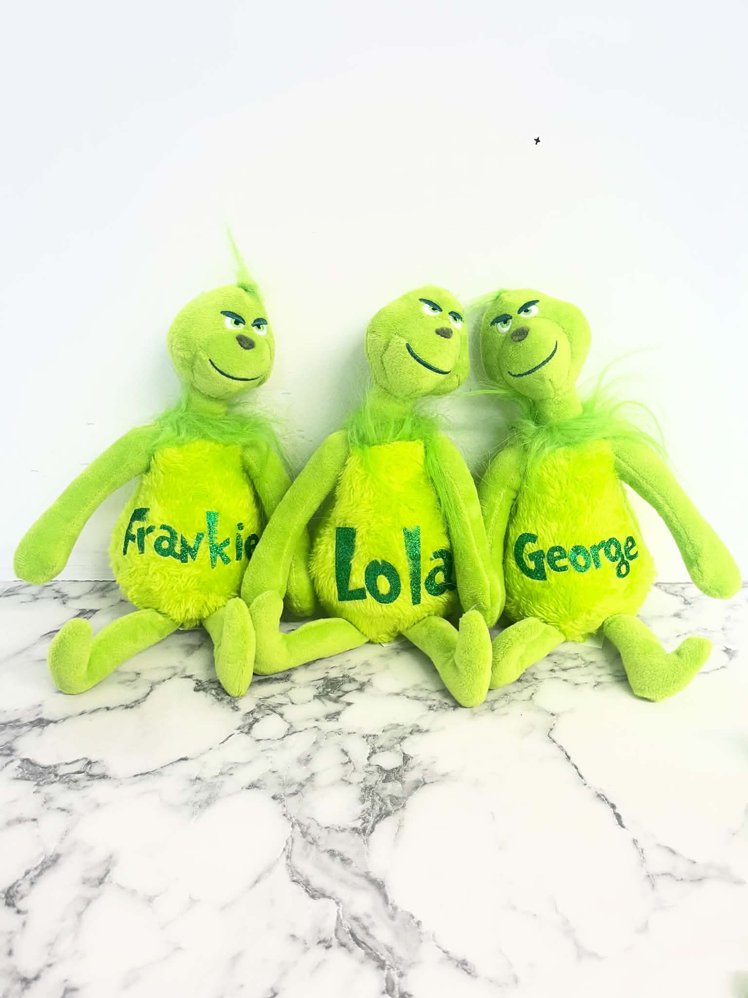 Three plush Grinch-style toys in bright green are arranged side by side on a white marble surface against a white wall. Each plush has a different name printed on the front: Frank, Lola, and George.