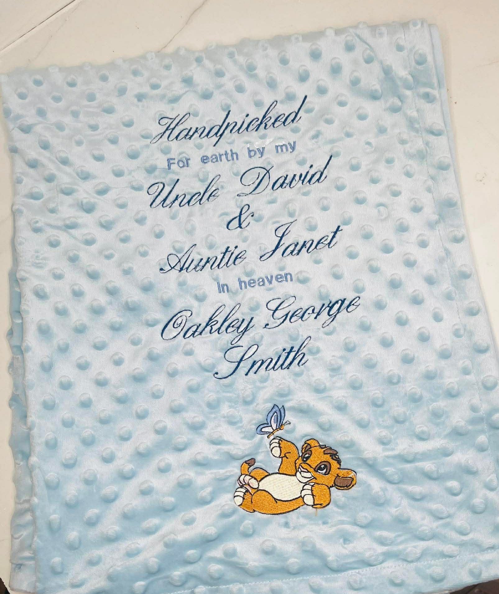 Blue baby blanket with embroidered text and a cartoon character on a white floor.