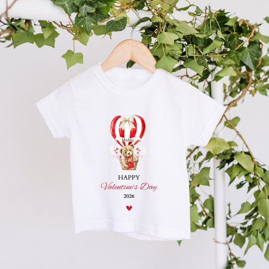 White t-shirt with Valentine's Day design featuring a teddy bear in a hot air balloon, hanging against a light background with greenery.