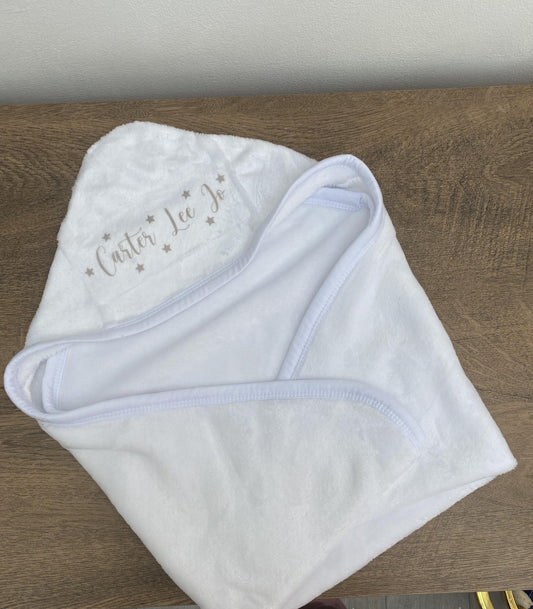 A white hooded baby blanket with personalized text embroidered on the side, displayed on a wooden surface.