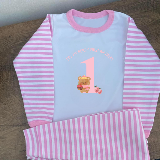 Pink and white striped children's pajama set with teddy bear and number 1 design on a wooden surface.