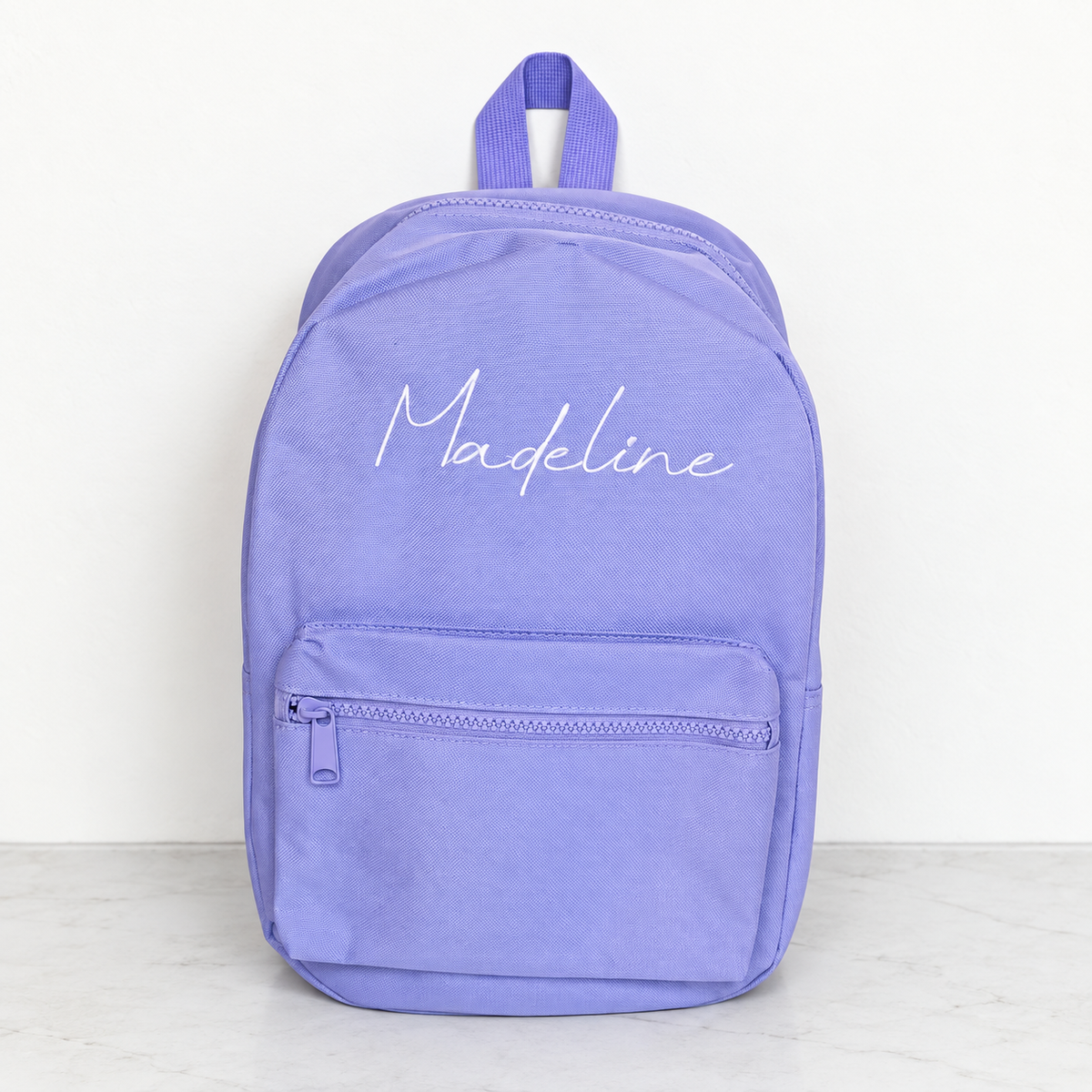 A lavender purple backpack with the name 'Madeline' embroidered in white script on the front, set against a plain white wall and light marble floor.