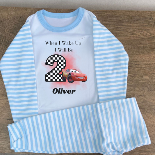 A personalized McQueen themed pyjama set for a child, with the pyjama top featuring a checkered pattern and the text 'When I wake up I will be 2' along with the McQueen logo. The pyjama bottoms are solid blue.