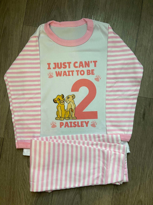 A personalised Lion King themed birthday pyjama set for a toddler, with 'I just can't wait to be 2' printed on the front, and a pair of pink striped shorts with a pattern on the side.