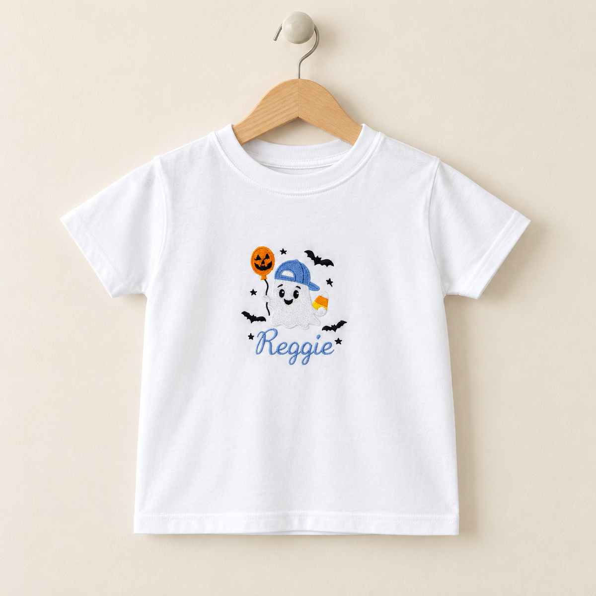 A white kids' short-sleeve t-shirt hanging on a wooden hanger against a light beige background. The shirt features a colorful graphic of a cartoon ghost wearing a blue cap, surrounded by bats, a jack-o'-lantern, and the name 'Reggie' in blue script.