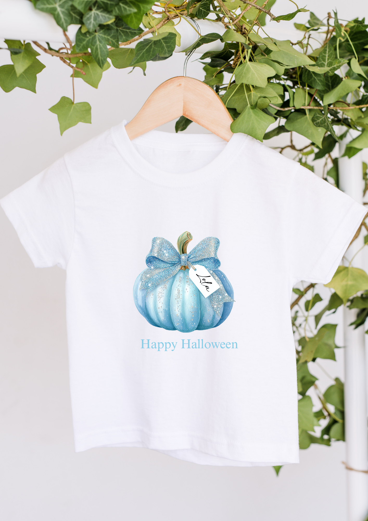 White t-shirt with blue pumpkin design and 'Happy Halloween' text on a hanger with greenery in the background.