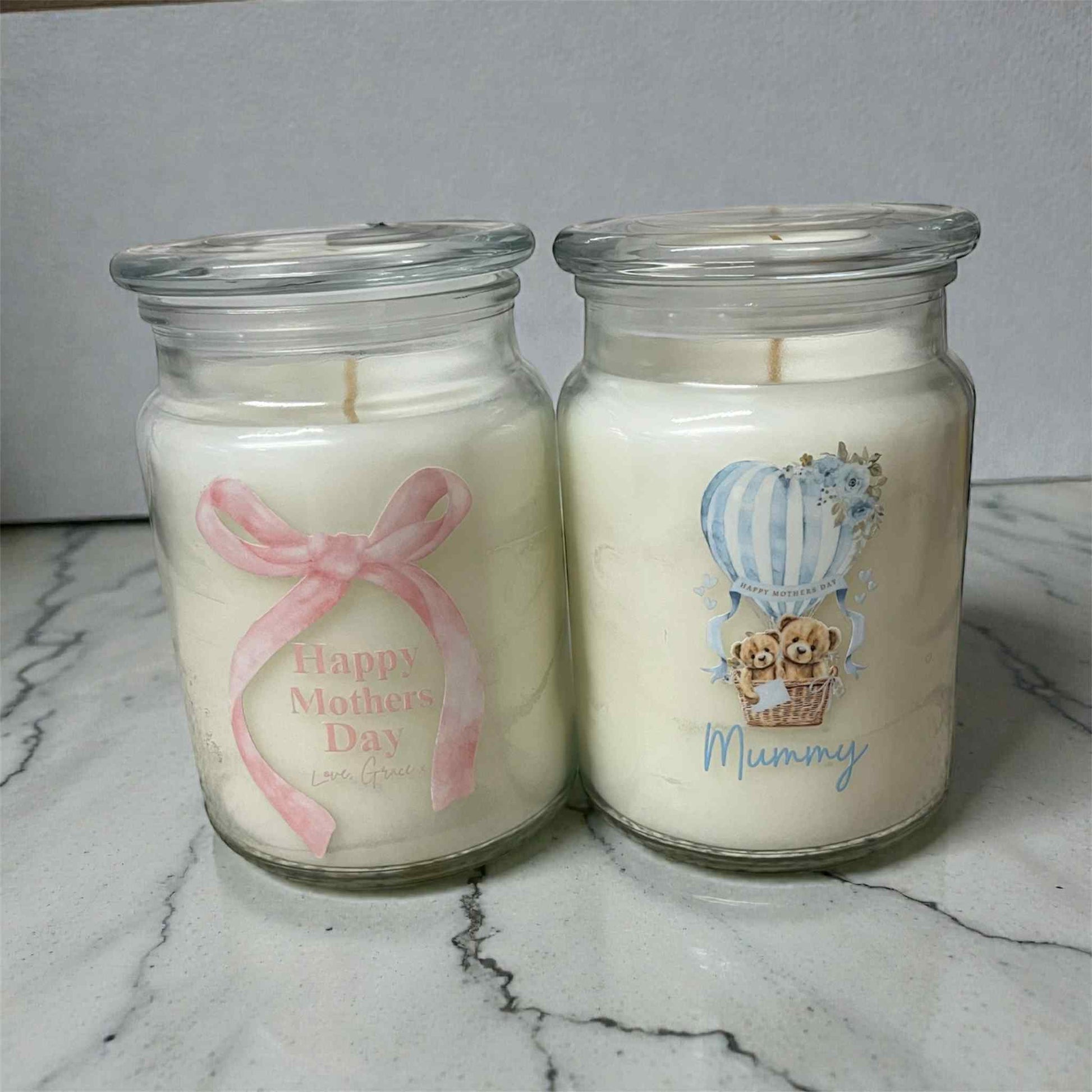 Two candles in glass jars with decorative labels on a marble surface.
