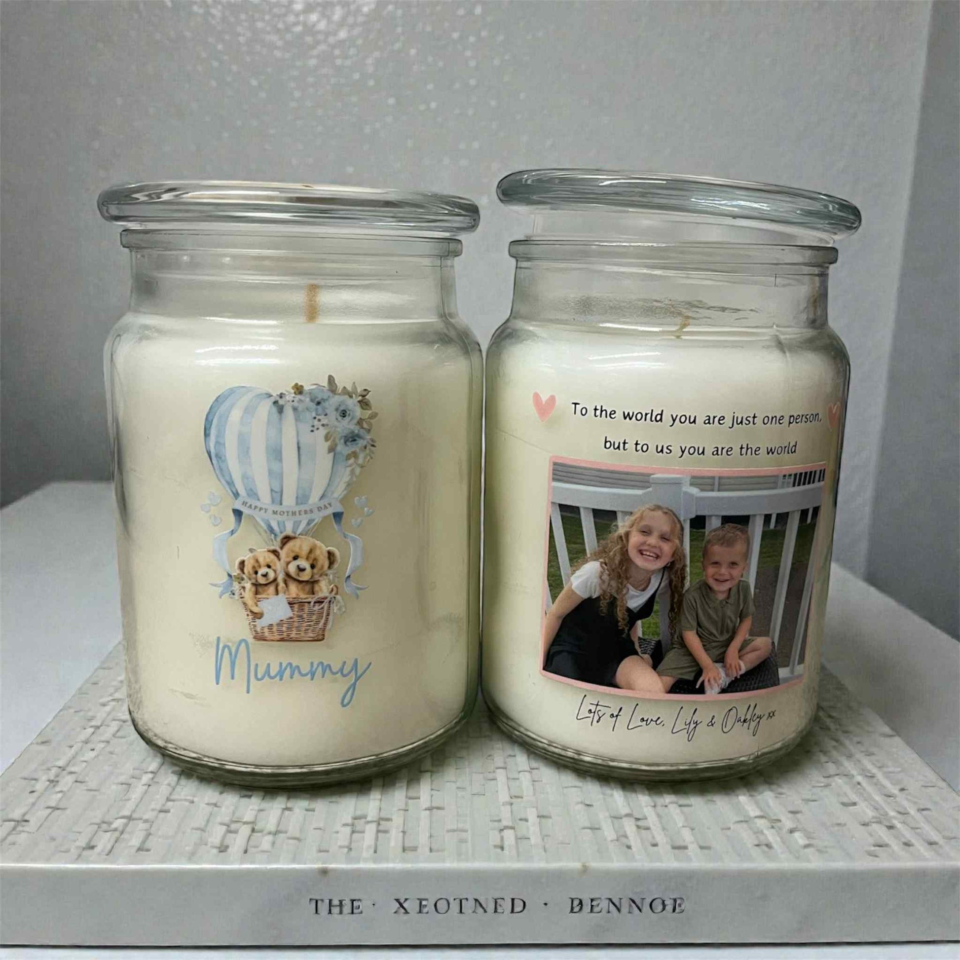 Two candles in glass jars with personalized designs and text on a textured surface.