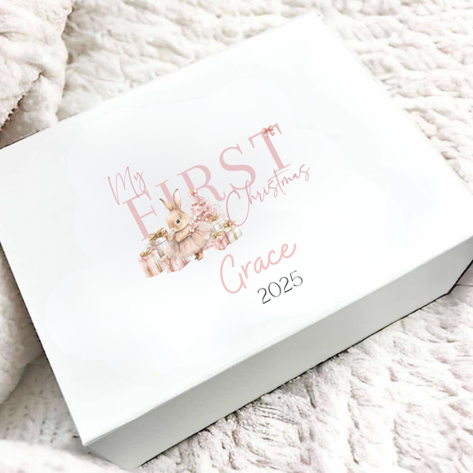 A white rectangular gift box with a custom printed design reading 'My FIRST Christmas Grace 2025' in pink script and a small bunny illustration, placed on a textured cream knit blanket.
