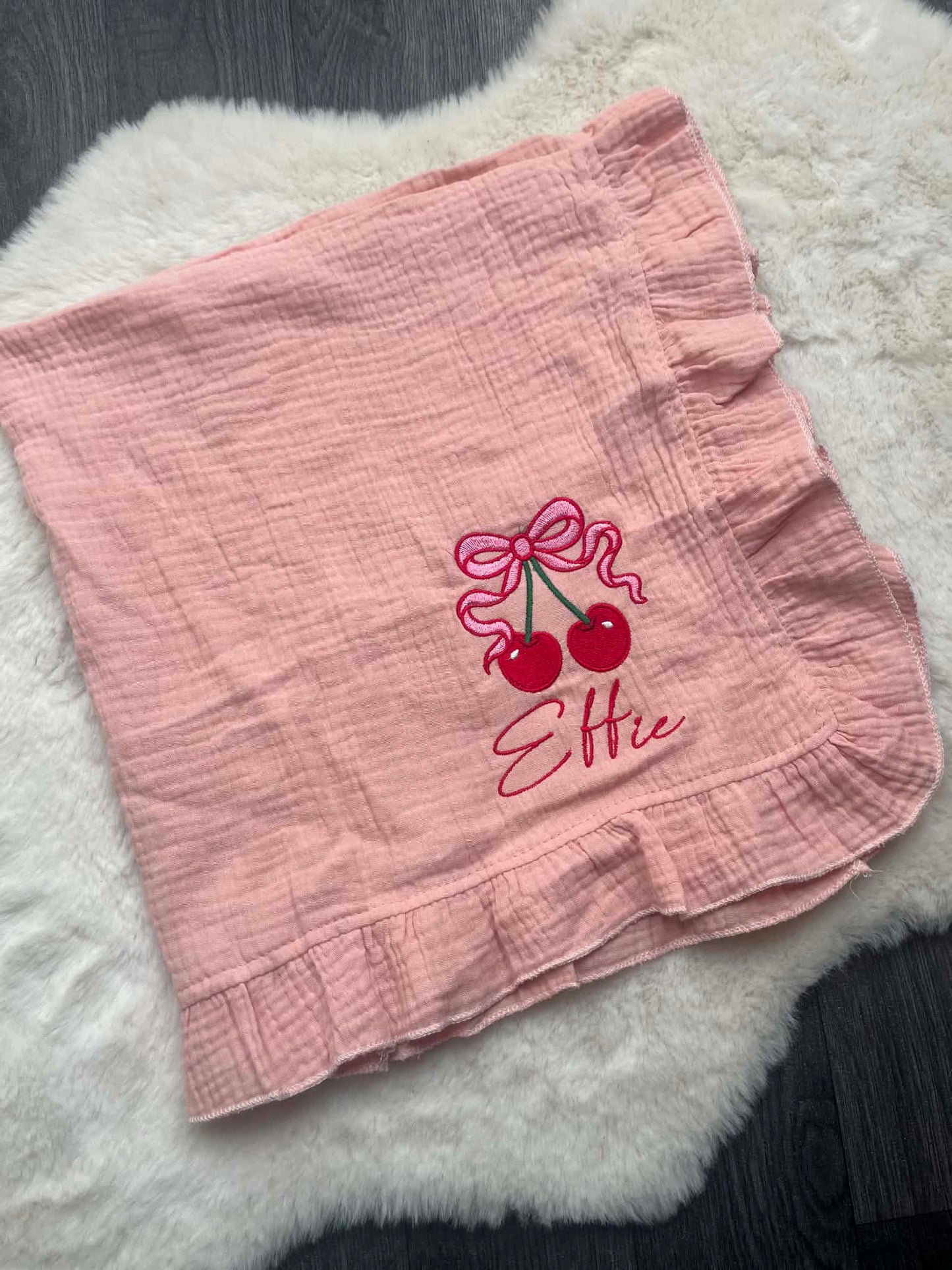 A pink crinkle gauze baby blanket with ruffled edges, featuring an embroidered cherry design and the name 'Ellie' in script. The blanket is laid flat on a white faux fur rug over a dark wood floor.
