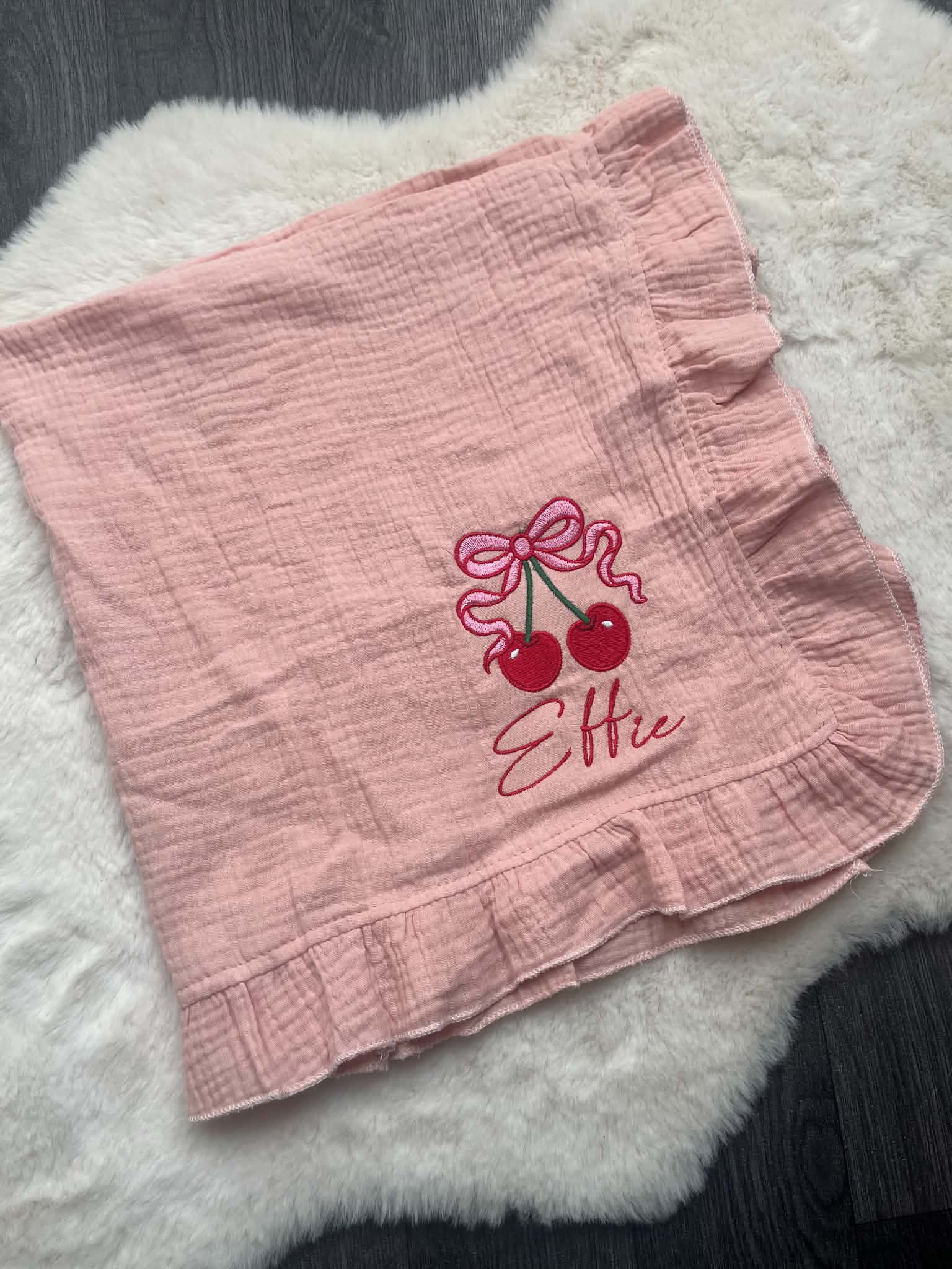 A pink crinkle gauze baby blanket with ruffled edges, featuring an embroidered cherry design and the name 'Ellie' in script. The blanket is laid flat on a white faux fur rug over a dark wood floor.