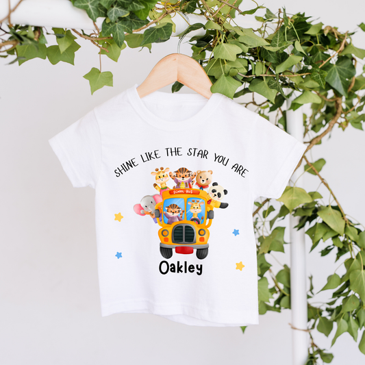 A white kids' t-shirt hanging on a wooden hanger from green ivy. The shirt features a colorful cartoon graphic of safari animals riding in a yellow school bus, with the text 'SHINE LIKE THE STAR YOU ARE' above and 'Oakley' below.