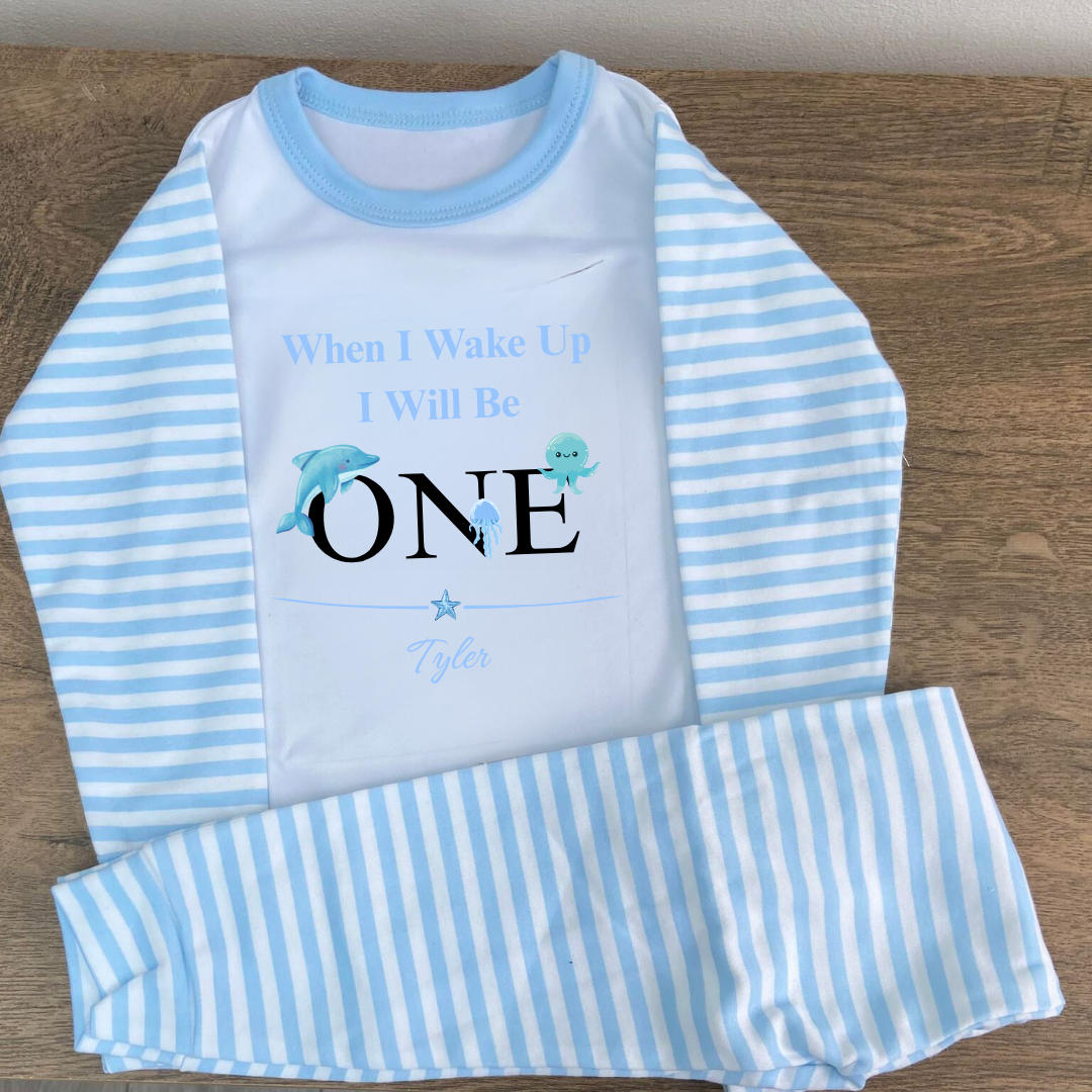 Children's blue and white striped pajama set with 'When I Wake Up I Will Be ONE' text on a wooden surface.