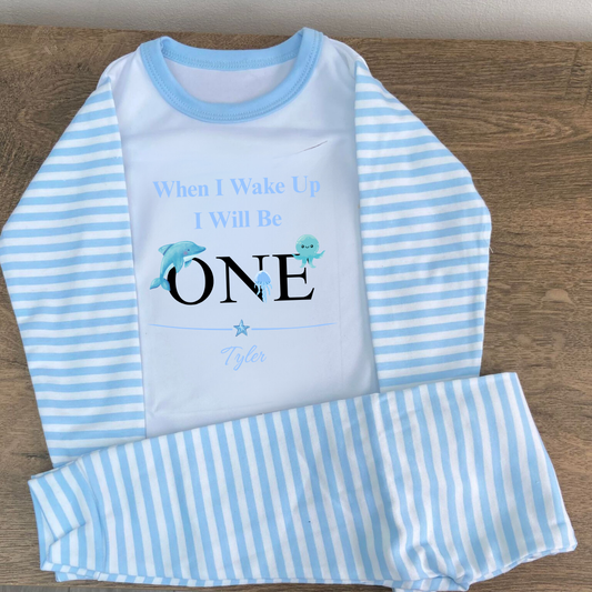 Children's blue and white striped pajama set with 'When I Wake Up I Will Be ONE' text on a wooden surface.