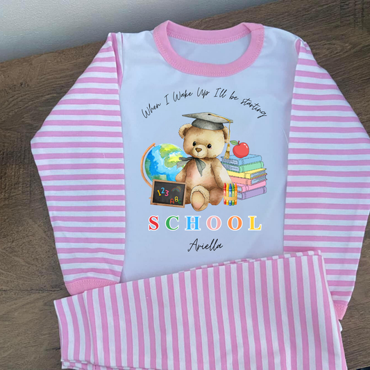 A children's long-sleeve pajama set laid flat on a wooden surface, featuring a white body with pink and white striped sleeves and matching striped pants. The front has a teddy bear school graphic with the text 'When I Wake Up I'll be starting SCHOOL' and the name 'Anella' in script.