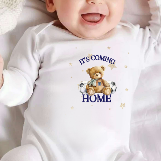 Baby wearing a white onesie with a teddy bear graphic and text 'It's Coming Home'.