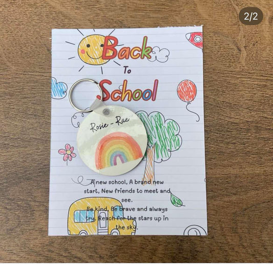 A personalized rainbow keyring with the text 'Back to School' and a drawing of a rainbow, sun, and school bus on a backing card.