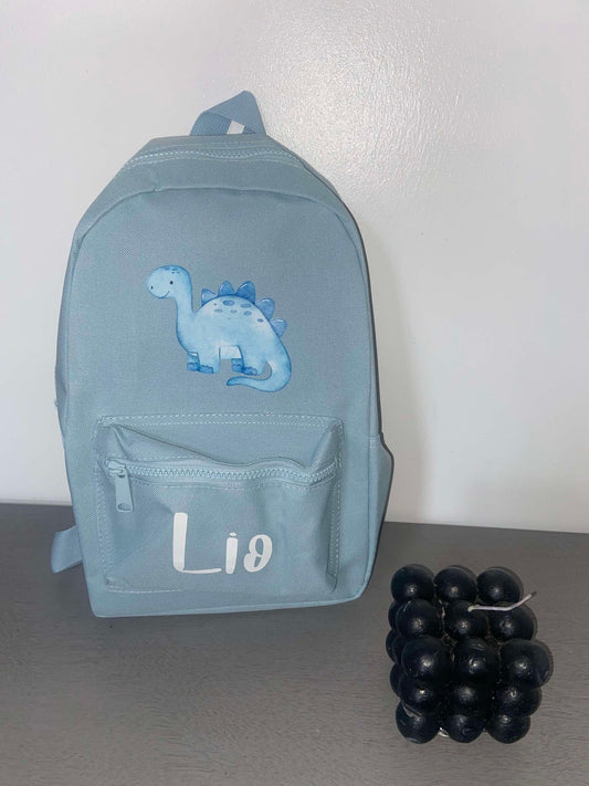 A blue personalised dinosaur-themed backpack with adjustable shoulder straps, displayed alongside a front pocket, on a surface with a few grapes.