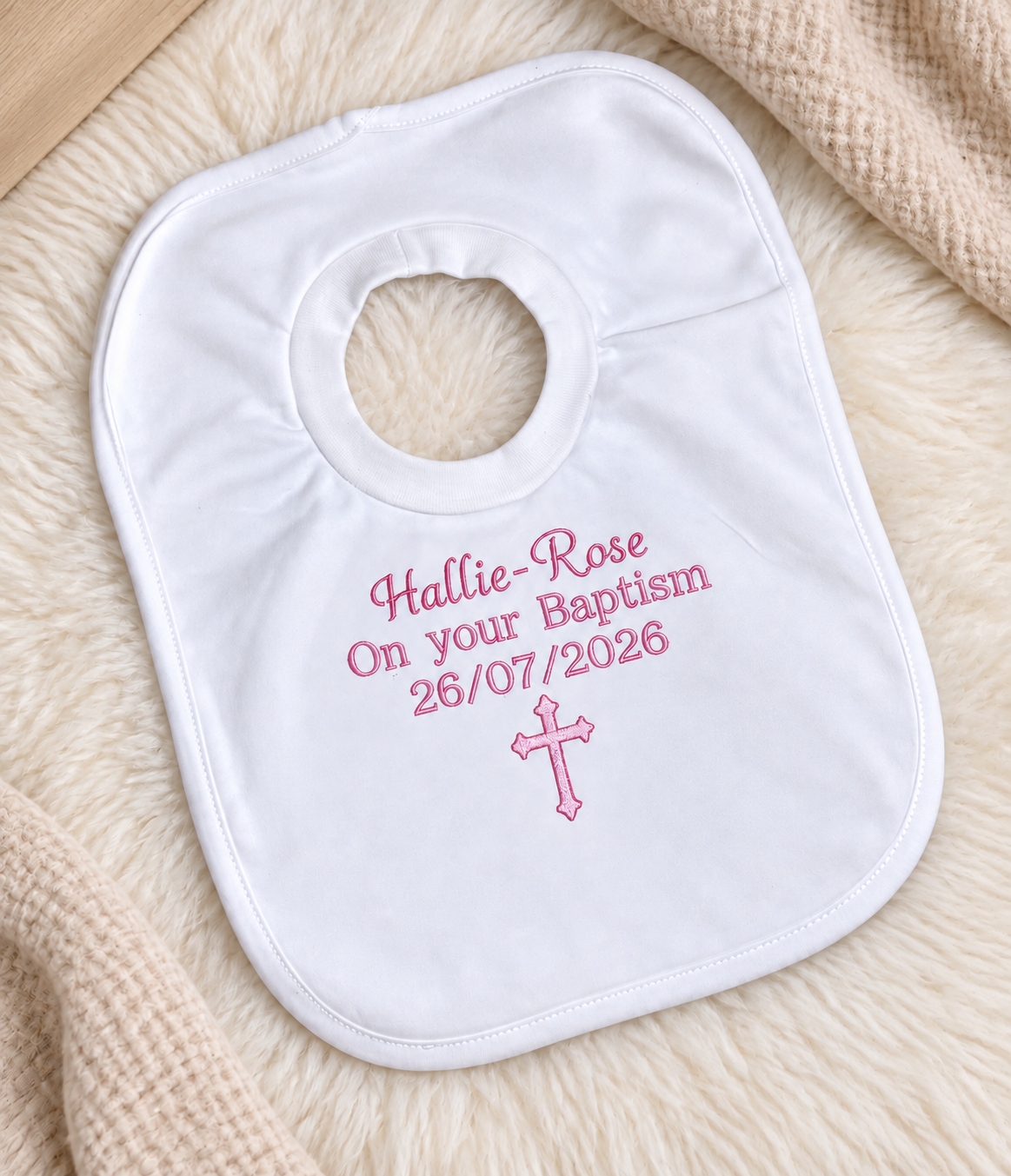 A white personalised baby bib with pink embroidered text reading 'Hallie-Rose On your Baptism 26/07/2026' and a pink cross, laid on a soft cream textured blanket.