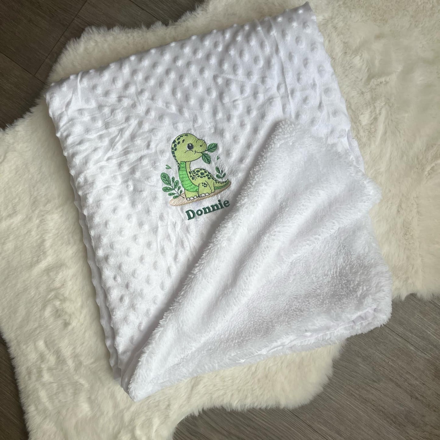 White baby blanket with a green dinosaur design and 'Donnie' text on a fluffy white surface.