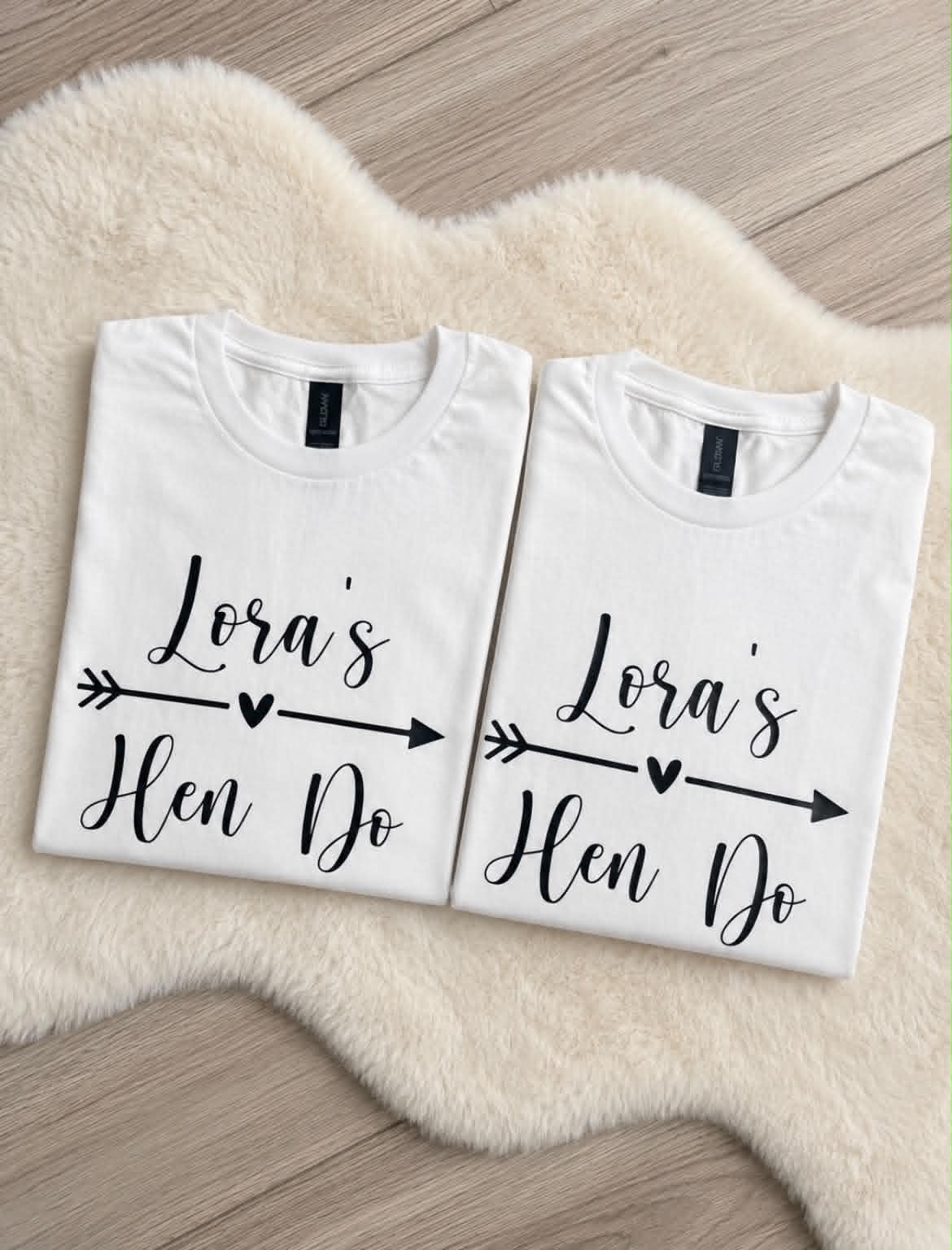 Two folded white graphic T-shirts displayed side by side on a cream faux-fur rug over a wood floor. Each shirt has black script text reading 'Lora's Hen Do' with an arrow and heart motif.