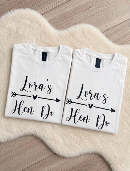 Two folded white graphic T-shirts displayed side by side on a cream faux-fur rug over a wood floor. Each shirt has black script text reading 'Lora's Hen Do' with an arrow and heart motif.