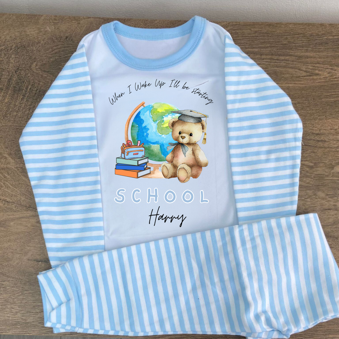 A children's long-sleeve pajama set laid flat on a wood surface, featuring light blue and white stripes on the sleeves and pants, with a white chest panel printed with a school-themed teddy bear illustration and the words 'When I Wake Up I'll be Fantasy SCHOOL Happy.'.