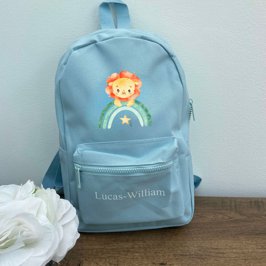 A light blue personalised backpack with a rainbow and a cartoon lion face on the front, with adjustable shoulder straps and a space for a name on the front.