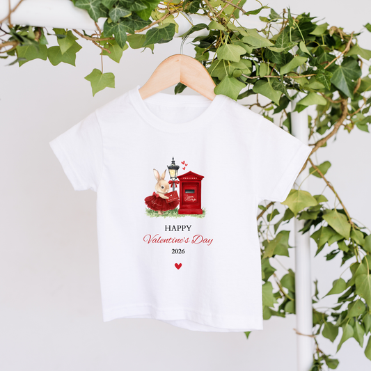 White t-shirt with Valentine's Day design featuring a rabbit and mailbox, hanging against a white background with green leaves.