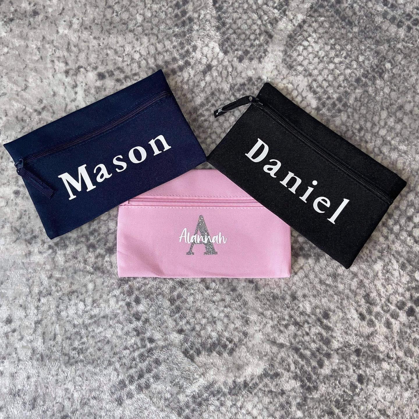 Customized pencil cases with different names printed on them, laid out on a textured surface.