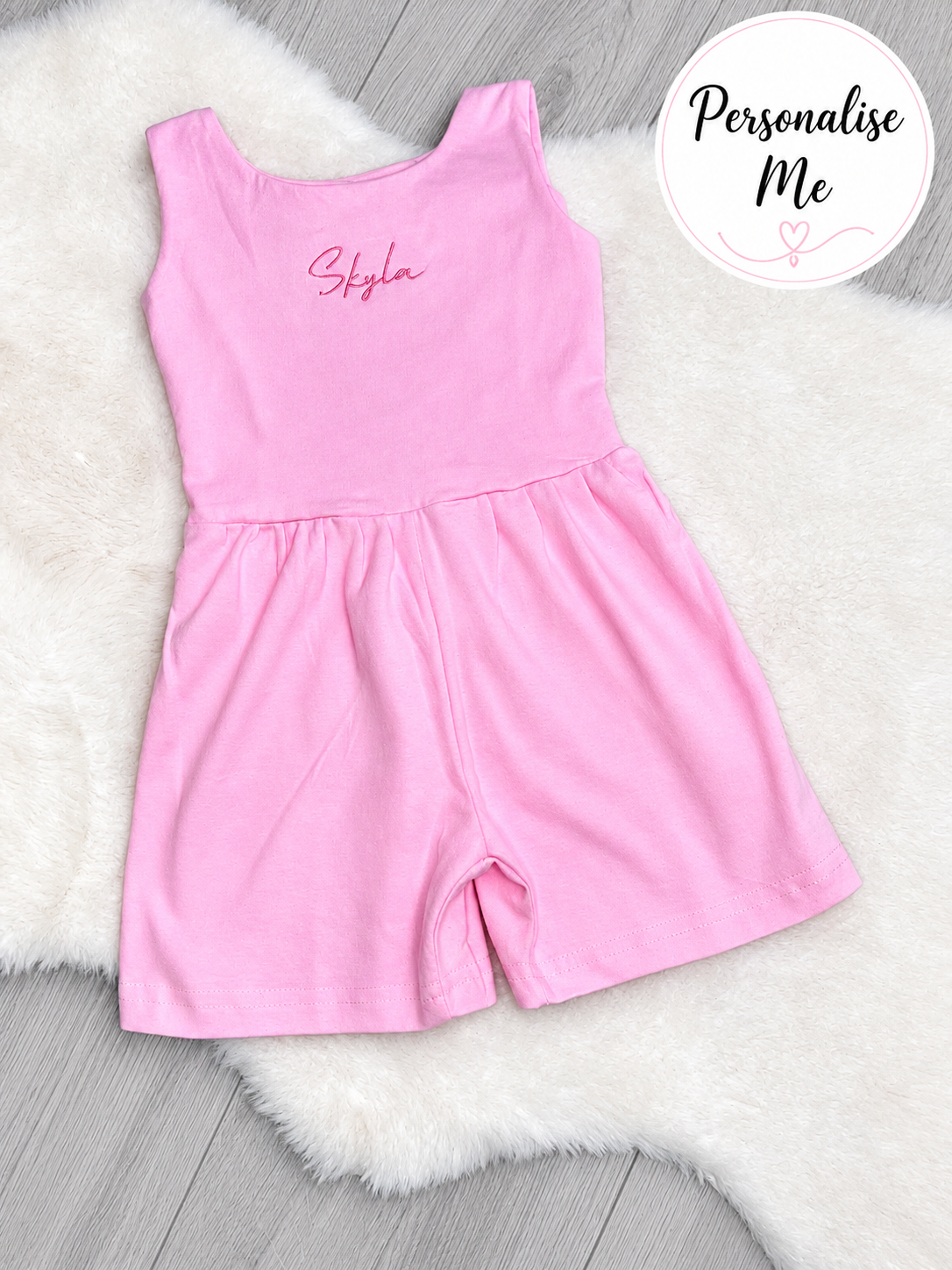 Personalised pink embroidered girls romper playsuit with custom name embroidery flat lay on white rug and grey laminate flooring.