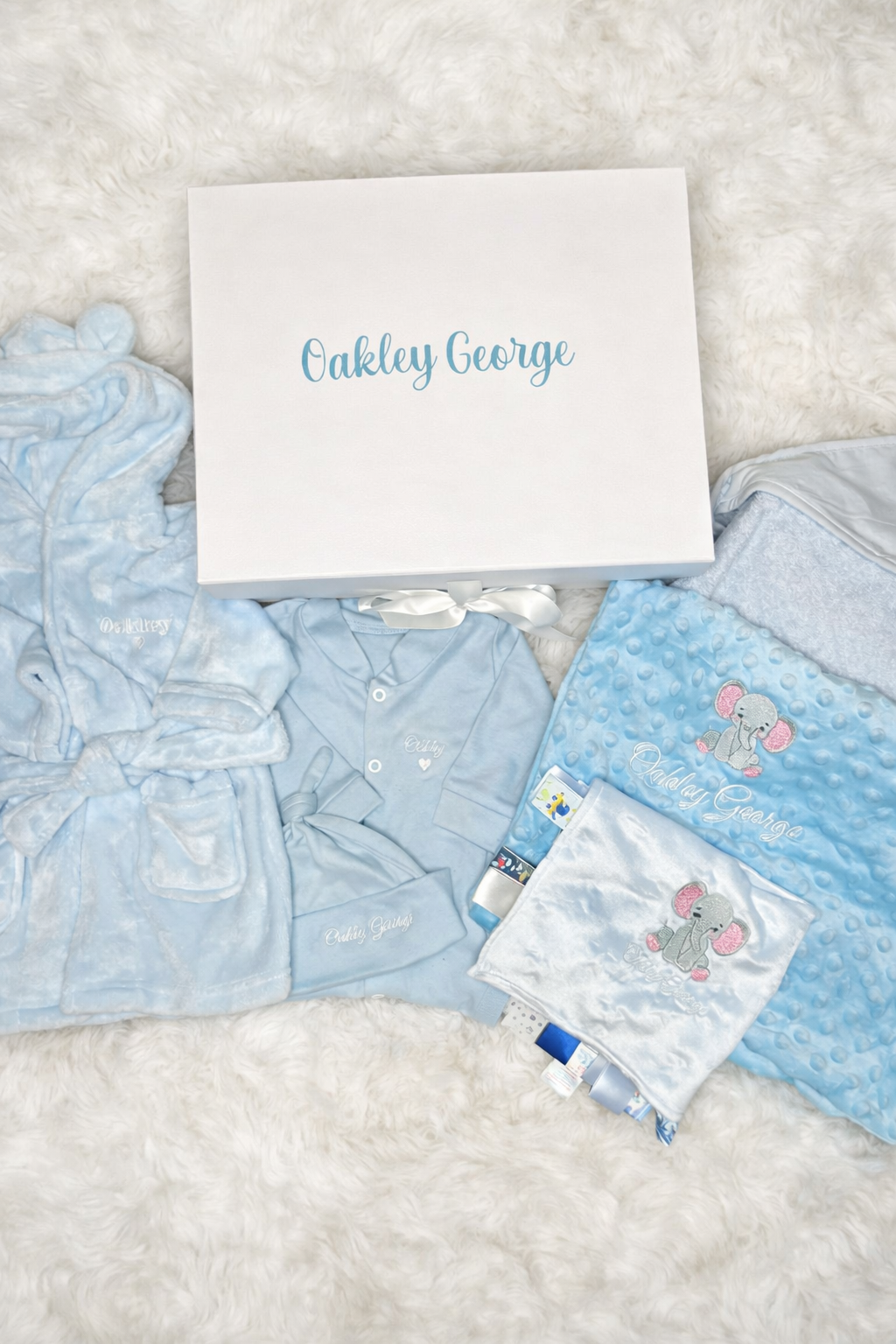 Baby boy clothing set with blue shirts and pants, white bib, and a white box labeled 'Oakley George' on a textured surface.