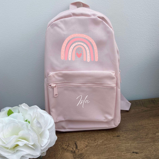 A pink personalised backpack for children with a rainbow design on the front and the name 'Lila' printed on the bottom.