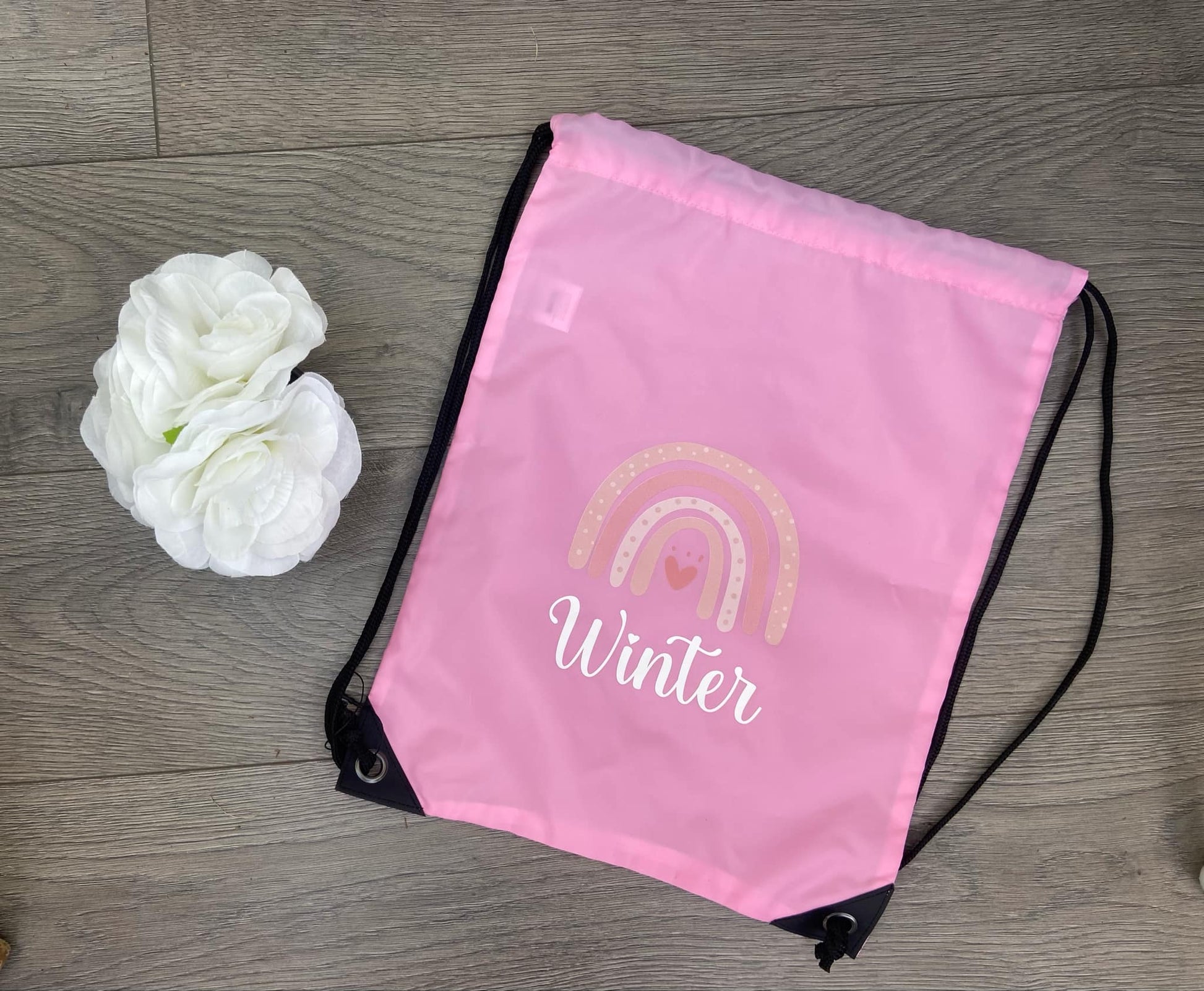 Pink drawstring bag with rainbow and 'Winter' text on a wooden surface with white flowers.