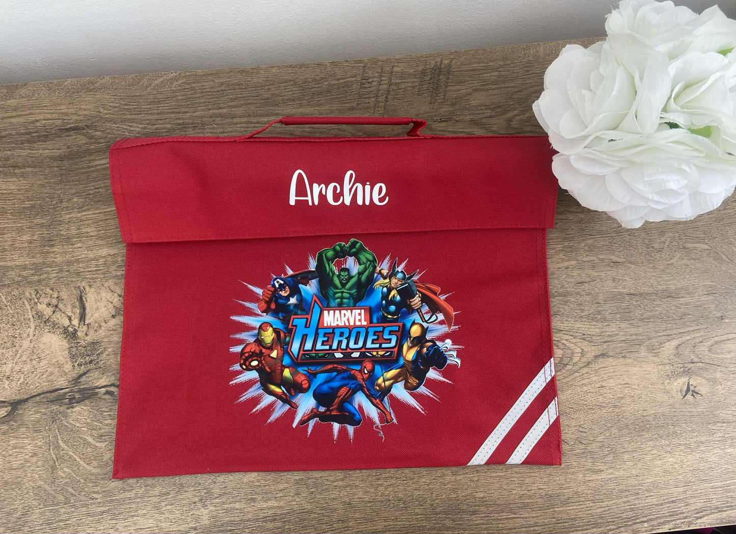 Red Book bag with Marvel Heroes design and 'Archie' text on a wooden surface with white flowers.