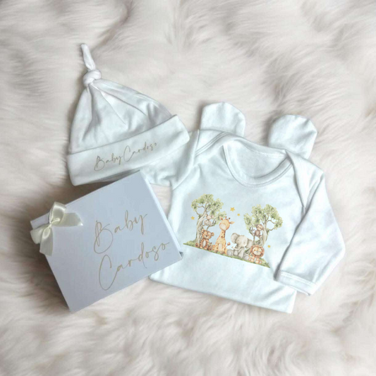 A baby gift set including a white hat, a white sleepsuit with a graphic design, and a personalized white box with a bow. The hat and sleepsuit have 'Baby Cardi' text on them, and the box is labeled 'Baby Cardi' as well.