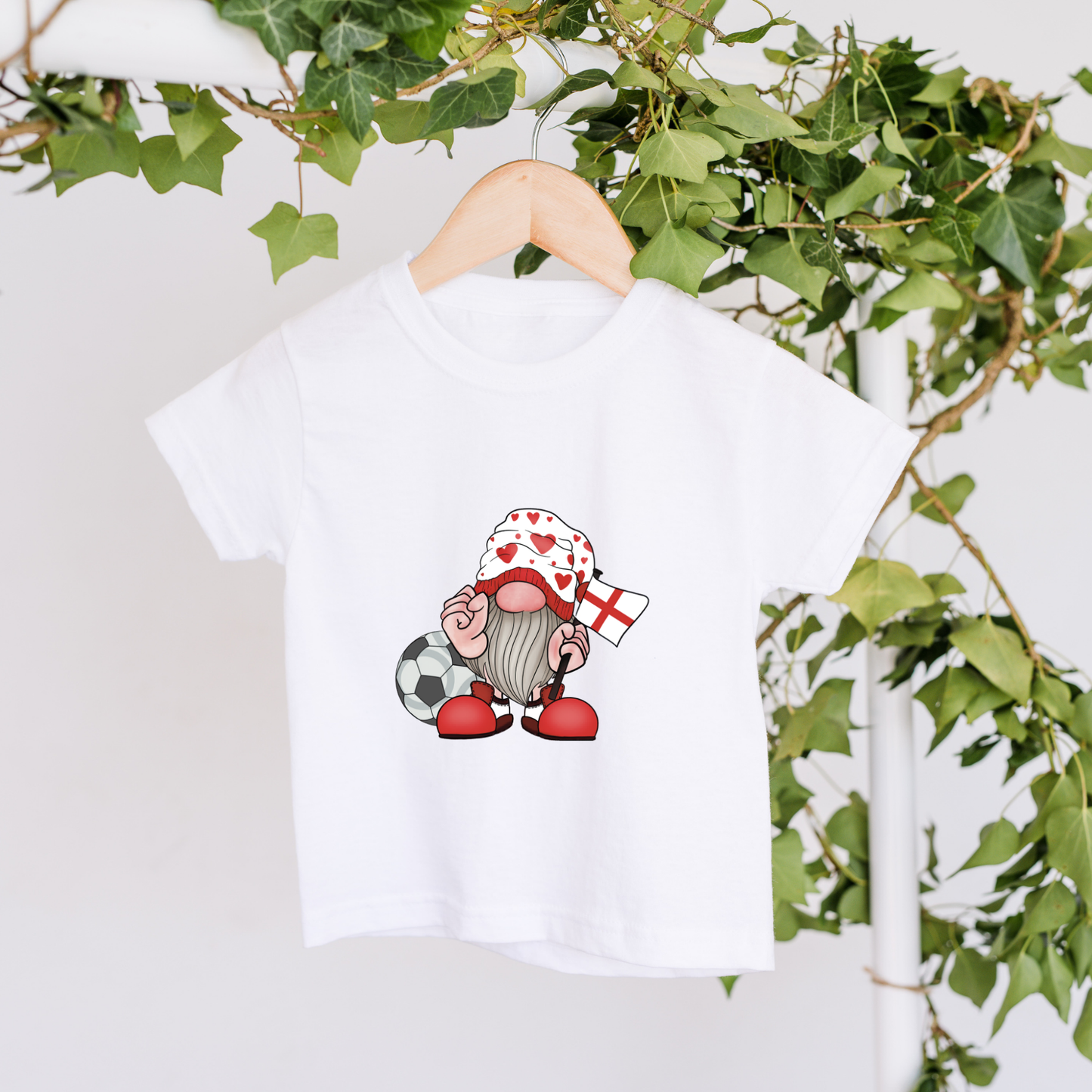 White t-shirt with a cartoon gnome design on a hanger against a white background with greenery.