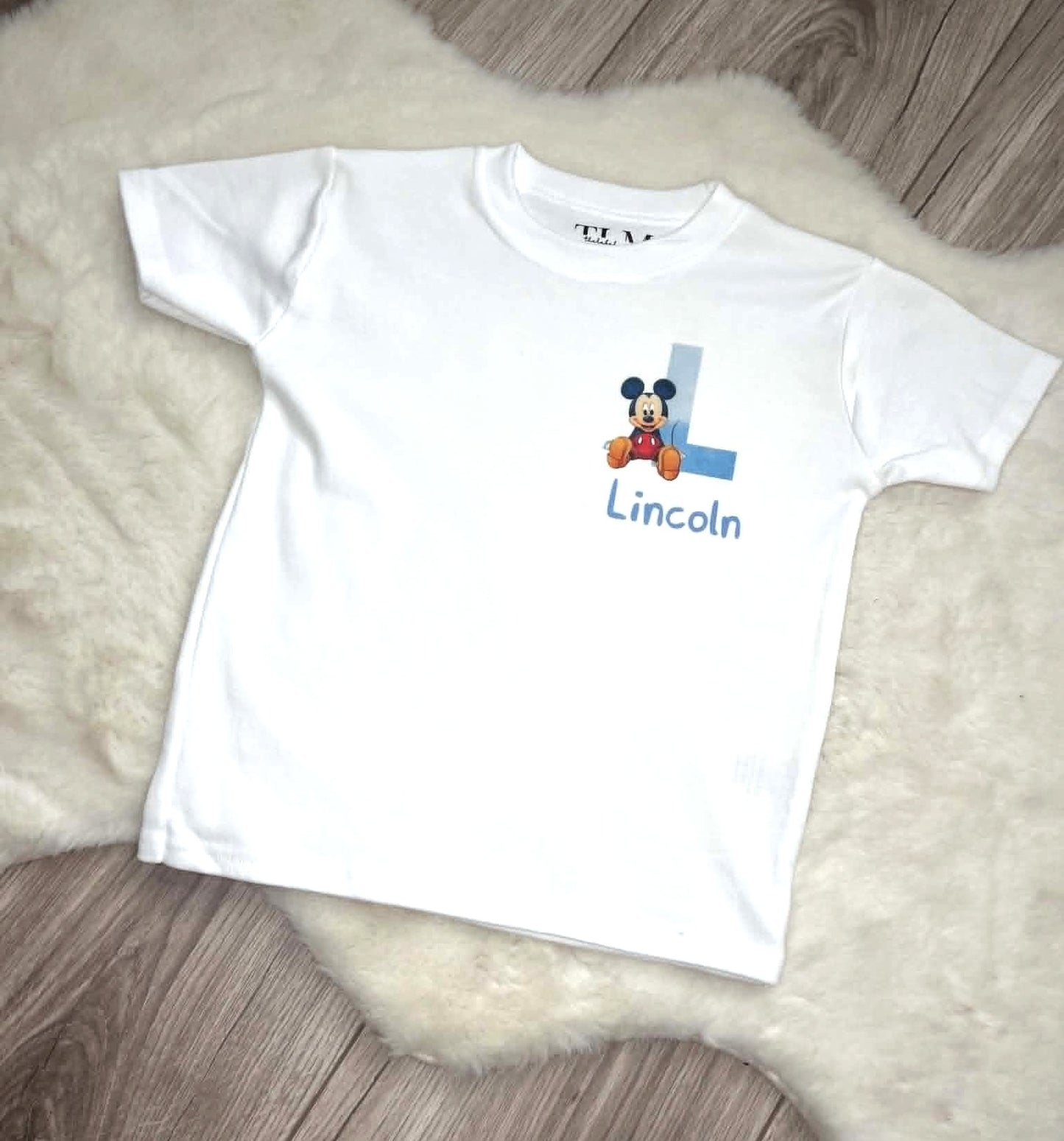 White personalised children’s T-shirt with blue initial, character design and custom name flat lay on white rug and grey laminate flooring.
