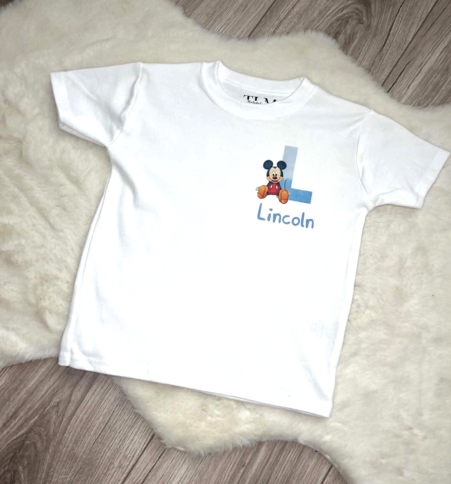 White personalised children’s T-shirt with blue initial, character design and custom name flat lay on white rug and grey laminate flooring.