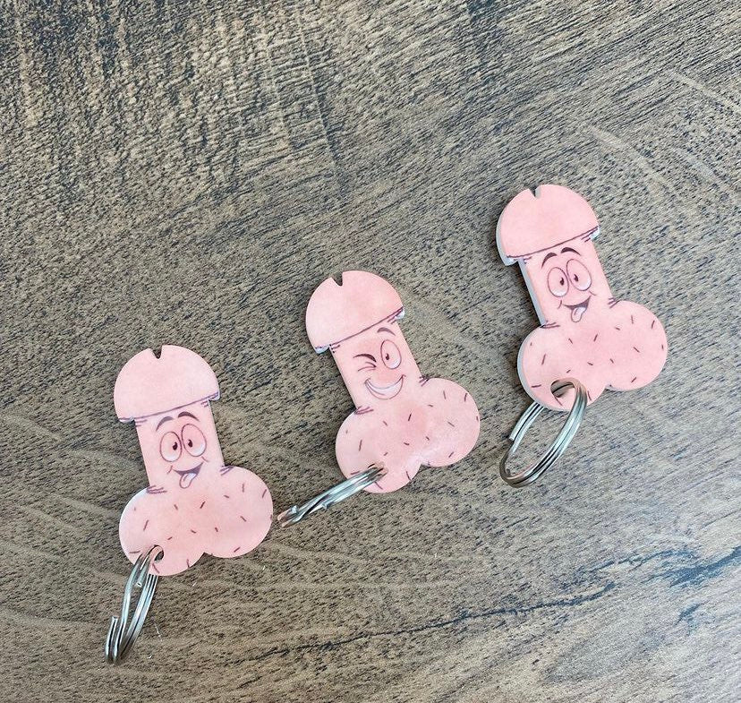 Three willy Tokens keychains shaped like cartoon characters with exaggerated features, placed on a wooden surface.