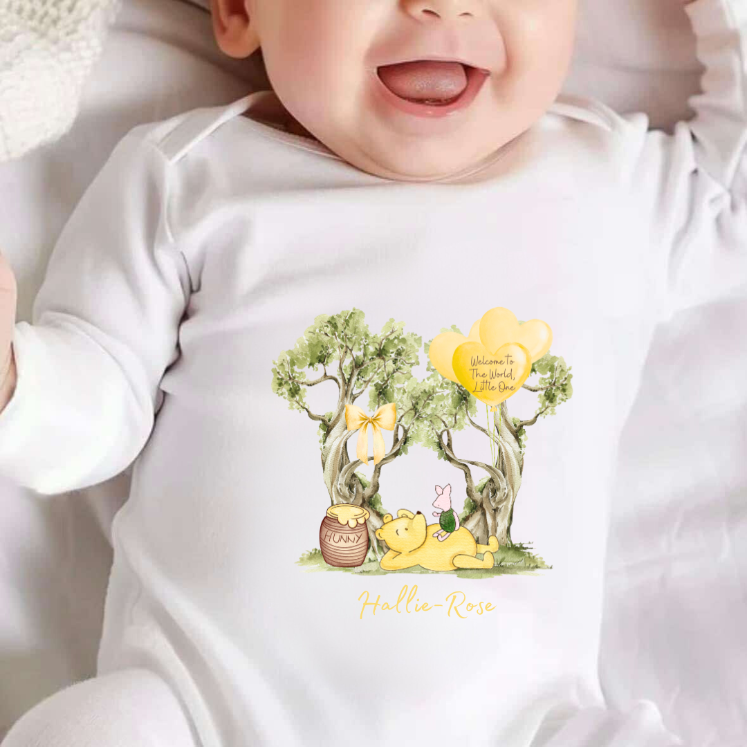 White sleepsuit with an image of pooh and piglet laid under a tree eating honey with the words welcome to the world