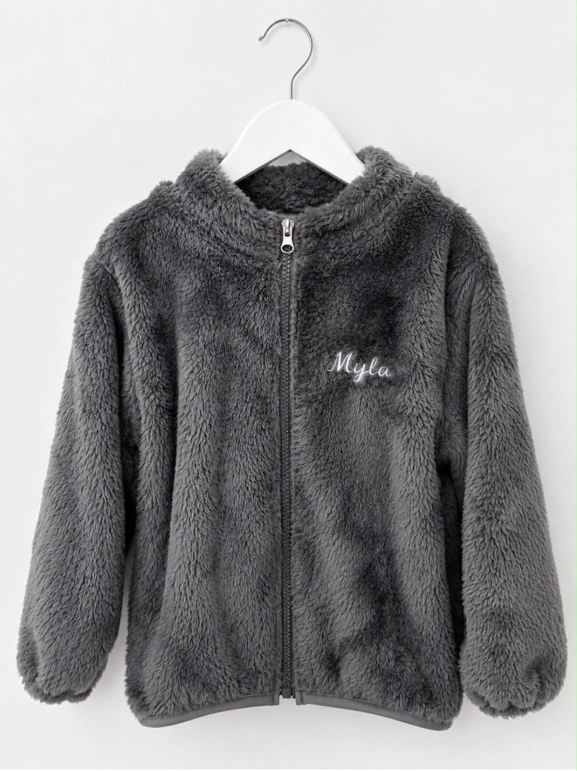 A dark gray plush faux-fur zip-up jacket with a hood, hanging on a white hanger against a light gray background. The word 'Myla' is embroidered in white script on the left chest.
