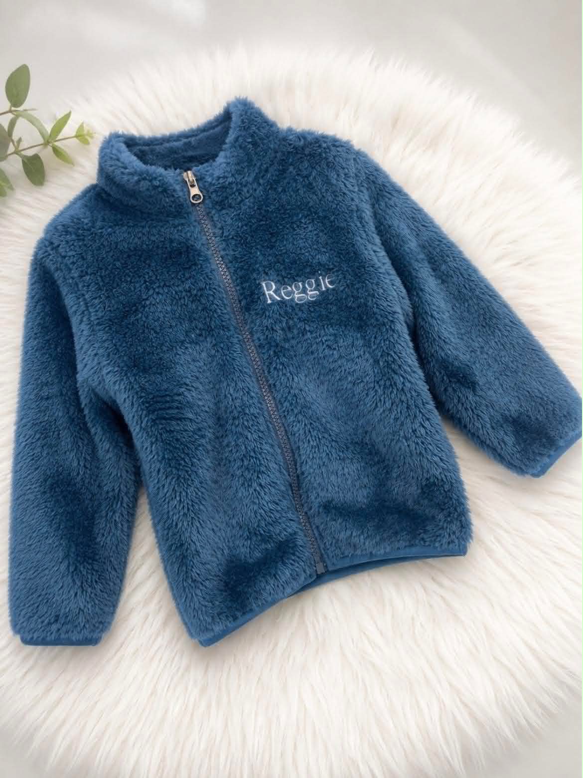 A blue sherpa fleece zip-up jacket with 'Reggie' embroidered on the chest, laid flat on a white faux fur surface.