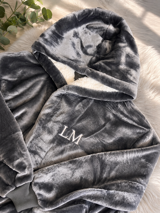 Gray plush blanket with embroidered initials on a light wooden surface.